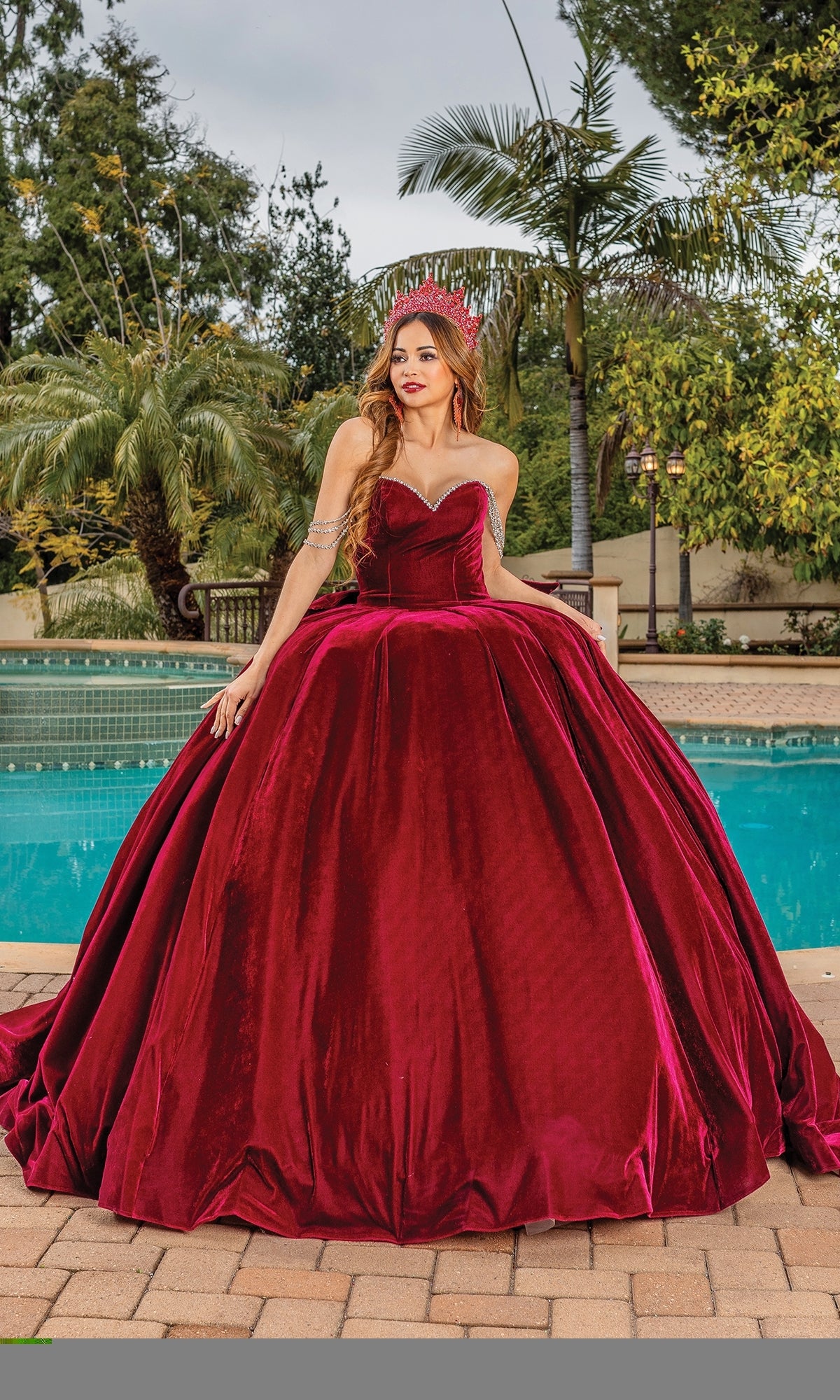 A woman in the Quinceanera Dress 1817 by Dancing Queen, a deep red velvet ball gown with a sweetheart strapless neckline and crown, stands by a swimming pool with trees and a fence in the background.